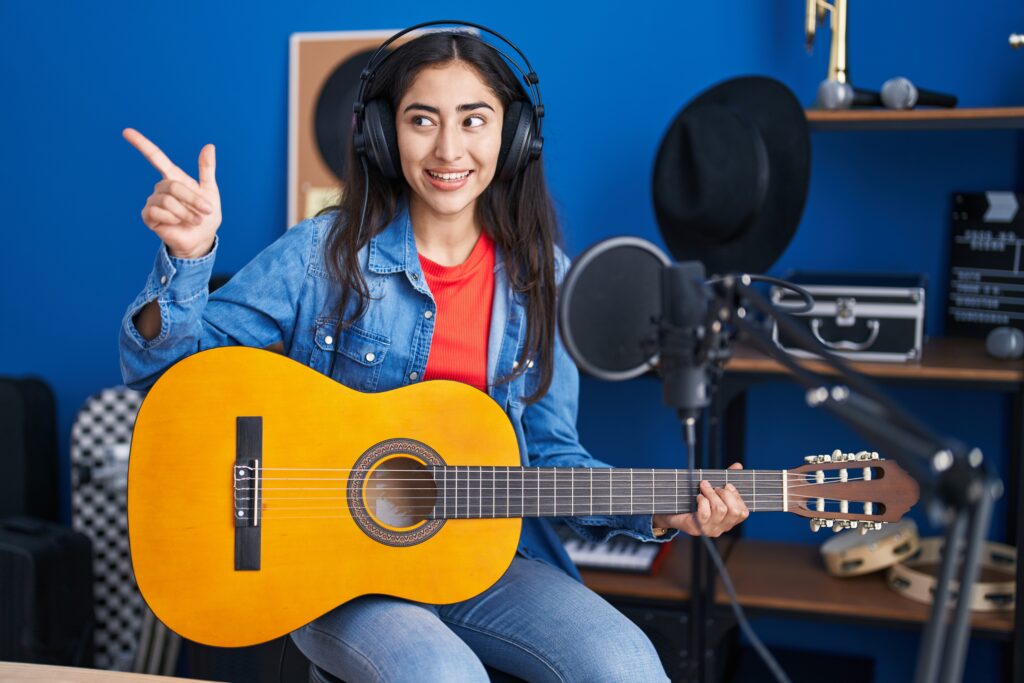 young teenager girl playing classic guitar at music studio pointing thumb up to the side smiling happy with open mouth